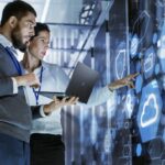 Two IT professionals in a data center review a holographic dashboard on a wall, laptop in hand, analyzing cloud and network data.